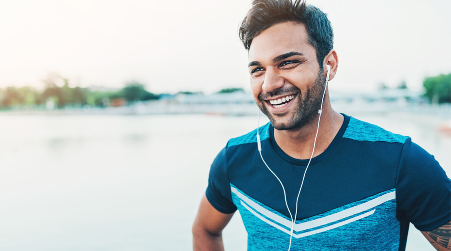 male in exercise shirt and headphones smiling with waterfront in background